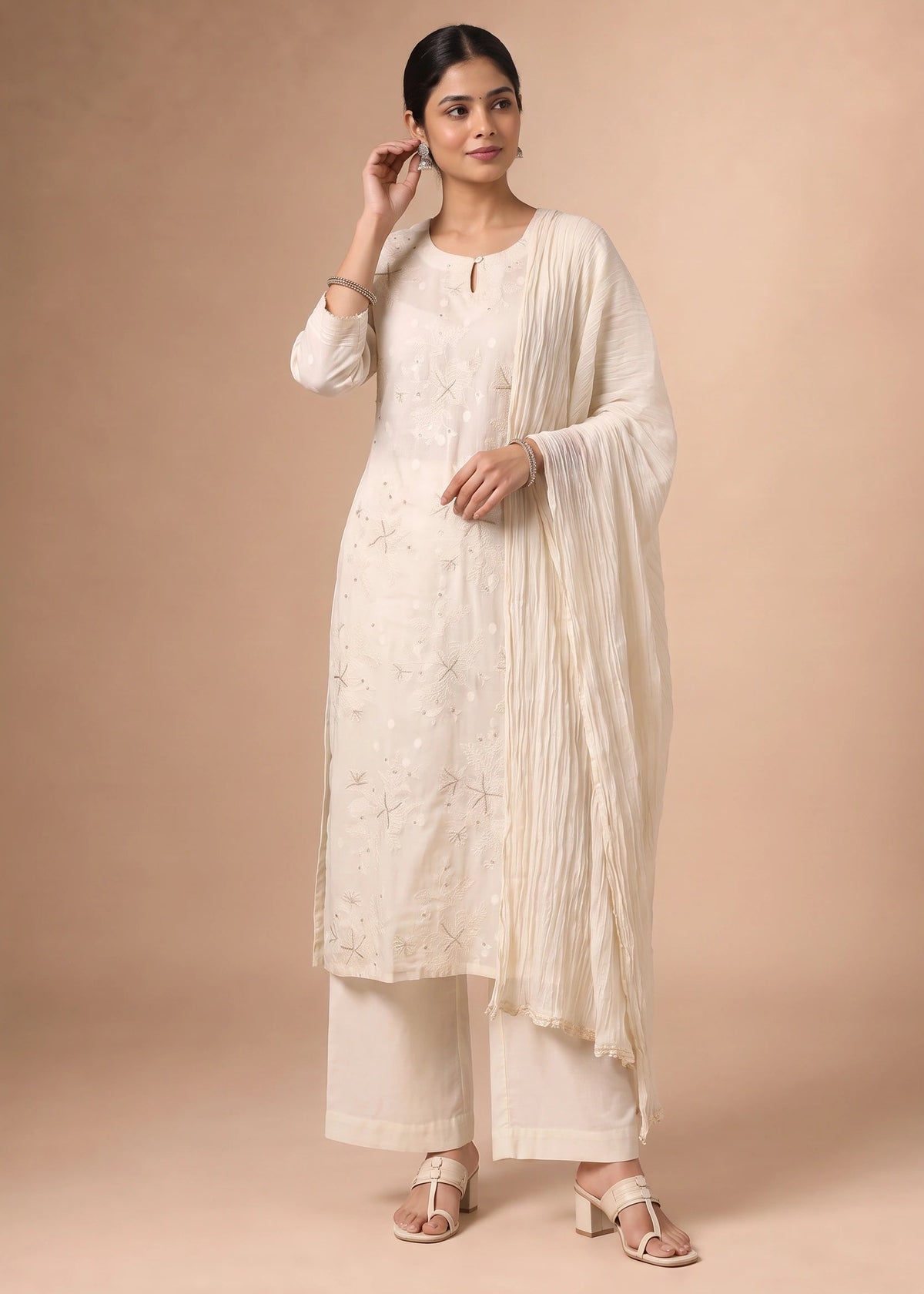 A woman modeling a traditional Indian outfit consisting of a cream-colored embroidered kurta, matching trousers, and a draped dupatta, set against a solid beige studio background.