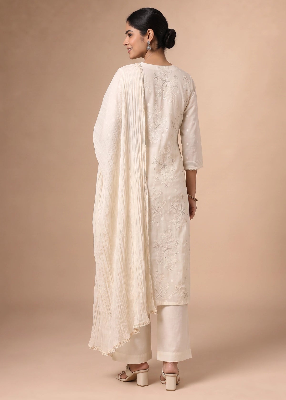 A woman modeling a traditional off-white/cream ethnic suit with floral embroidery, featuring a long kurta, matching pants, and a draped dupatta, shown from the back against a solid beige studio backdrop.
