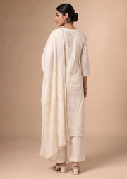 A woman modeling a traditional off-white/cream ethnic suit with floral embroidery, featuring a long kurta, matching pants, and a draped dupatta, shown from the back against a solid beige studio backdrop.