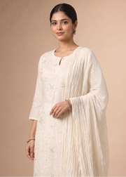 A woman modeling a traditional off-white/cream ethnic kurta set with a matching pleated dupatta, set against a solid beige studio background.