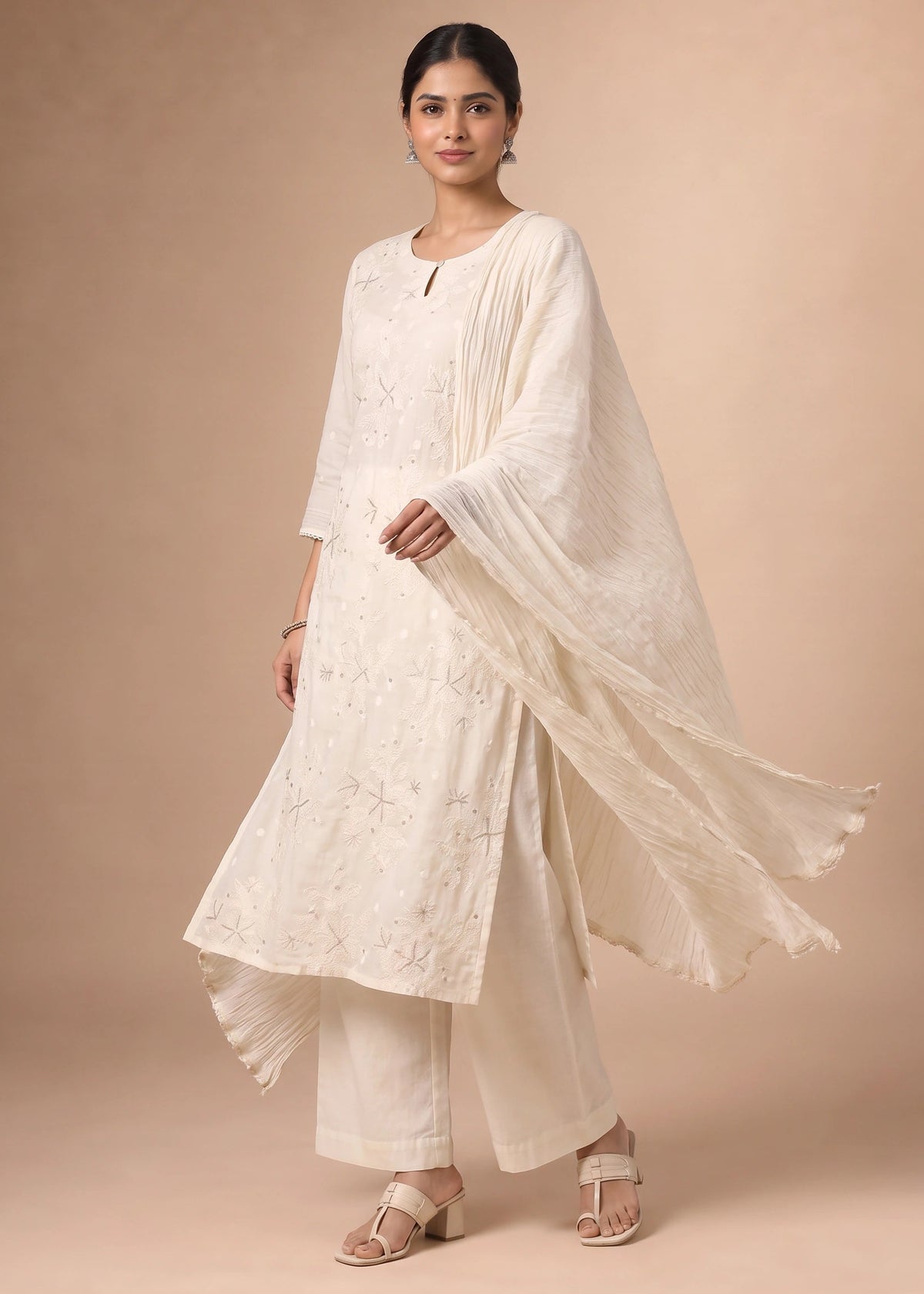 A woman modeling a traditional white embroidered kurta set with matching wide-leg pants and a flowing dupatta, posed against a solid beige studio backdrop.