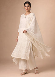 A woman modeling a traditional white embroidered kurta set with matching wide-leg pants and a flowing dupatta, posed against a solid beige studio backdrop.