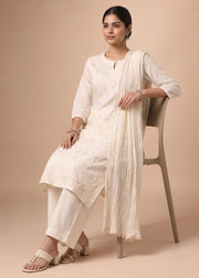 A woman seated on a chair wearing an off-white embroidered kurta with matching pants and a draped pleated dupatta, photographed against a plain beige studio background.