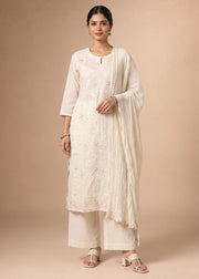 A woman modeling a traditional Indian outfit consisting of an off-white embroidered kurta, matching trousers, and a draped dupatta, set against a solid beige studio background.