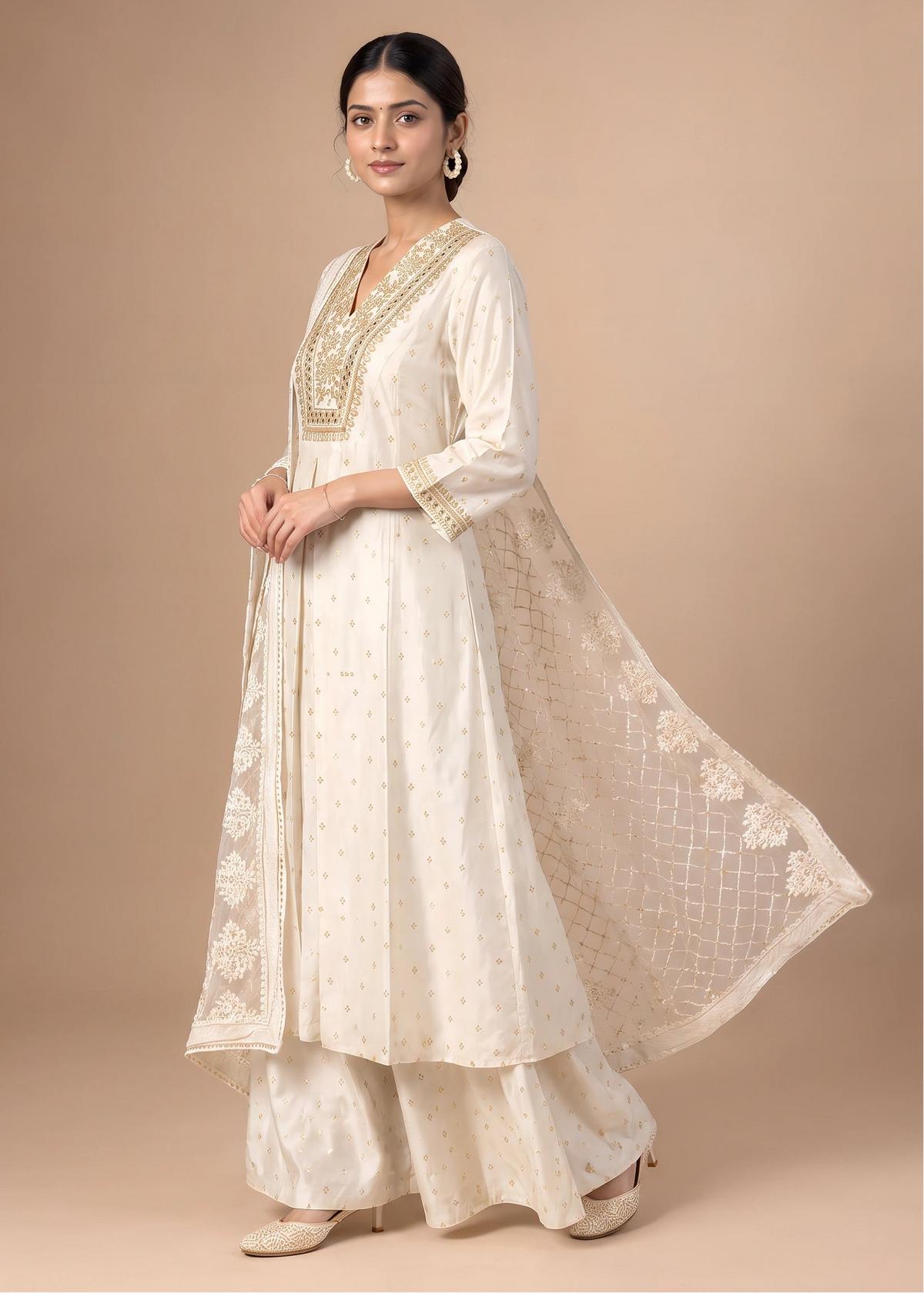 A woman models a traditional Indian outfit consisting of an off-white Anarkali kurta with gold embroidery, matching wide-leg pants, and a sheer embroidered dupatta, set against a solid beige studio background.