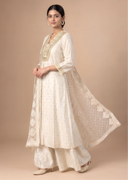 A woman models a traditional Indian outfit consisting of an off-white Anarkali kurta with gold embroidery, matching wide-leg pants, and a sheer embroidered dupatta, set against a solid beige studio background.