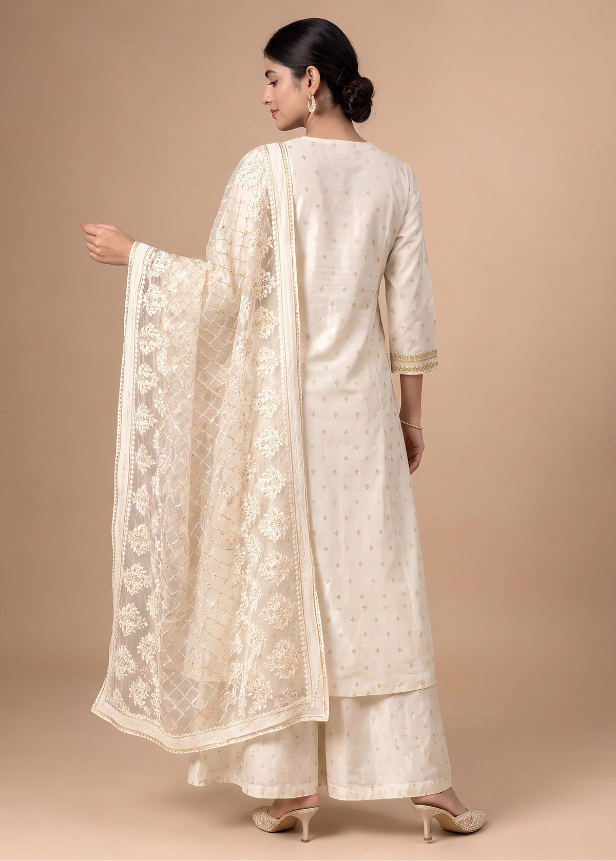 A woman modeling a traditional Indian salwar kameez suit in cream with gold dot embroidery and a matching embroidered dupatta, shown from the back against a solid beige studio background.