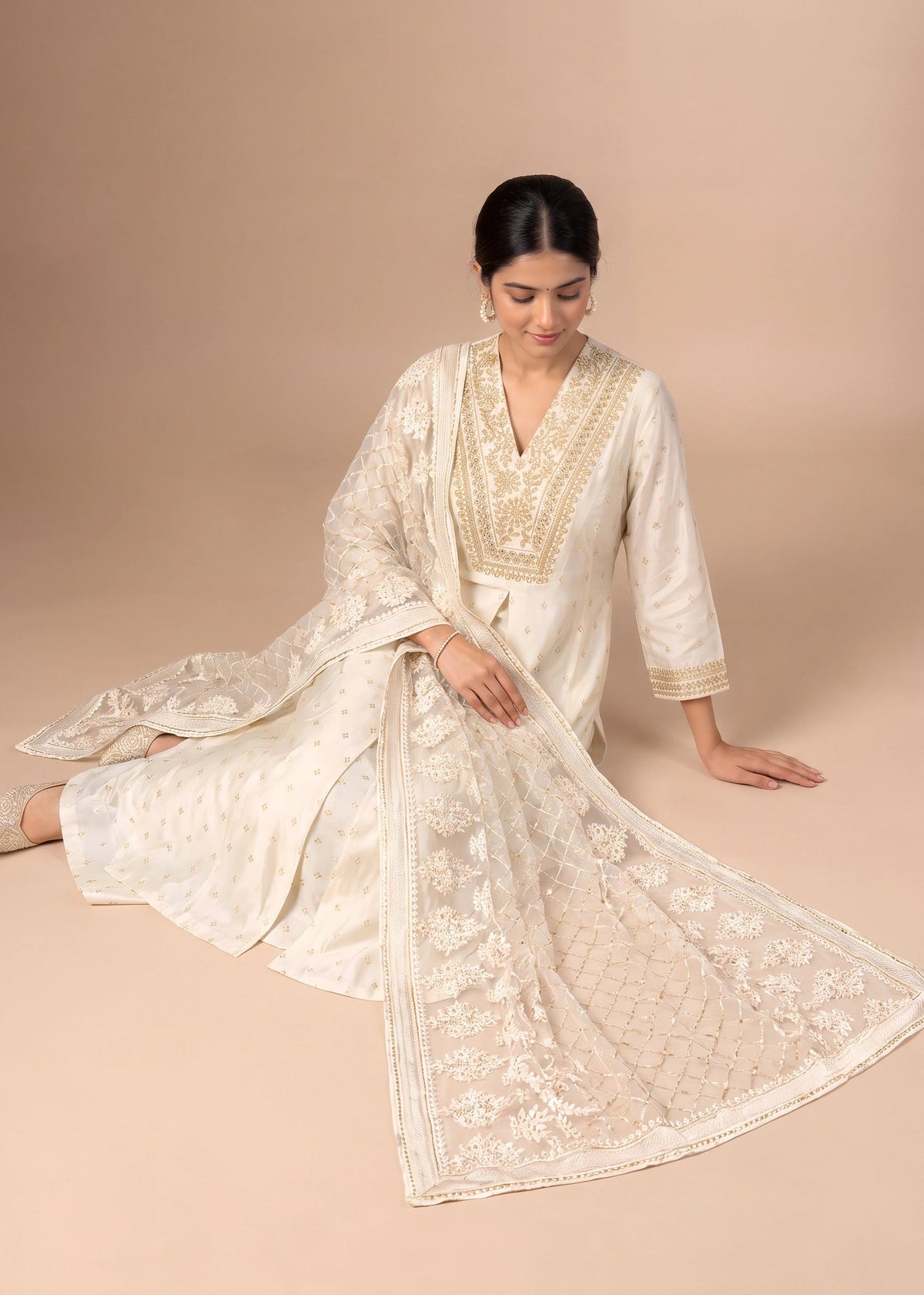 A woman models a traditional white Indian salwar kameez suit with intricate gold embroidery and a matching sheer dupatta, posed against a solid beige studio backdrop.