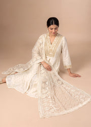A woman models a traditional white Indian salwar kameez suit with intricate gold embroidery and a matching sheer dupatta, posed against a solid beige studio backdrop.