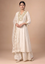 A woman modeling a traditional cream-colored Indian outfit, featuring a kurta with gold embroidery, matching palazzo pants, and a dupatta, set against a solid beige studio background.