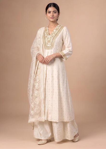 A woman modeling a traditional cream-colored Indian outfit, featuring a kurta with gold embroidery, matching palazzo pants, and a dupatta, set against a solid beige studio background.