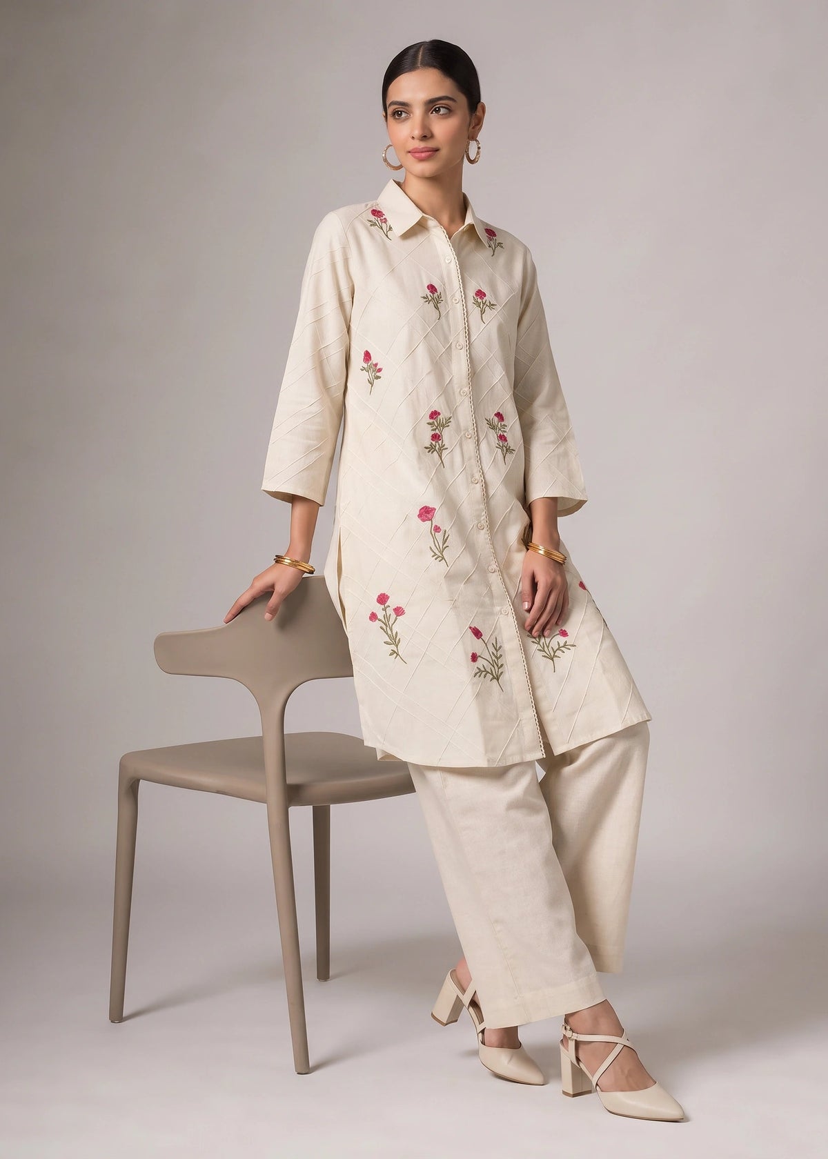 A woman models a cream-colored embroidered kurta and matching wide-leg trousers, leaning against a minimalist beige chair in a studio setting.