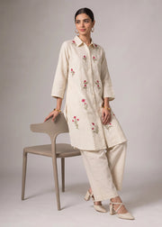 A woman models a cream-colored embroidered kurta and matching wide-leg trousers, leaning against a minimalist beige chair in a studio setting.