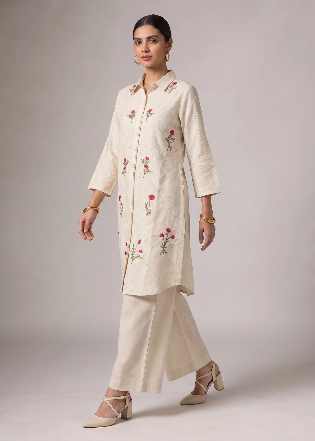 A woman models a cream-colored kurta and wide-leg trousers set with pink floral embroidery, set against a neutral studio background.