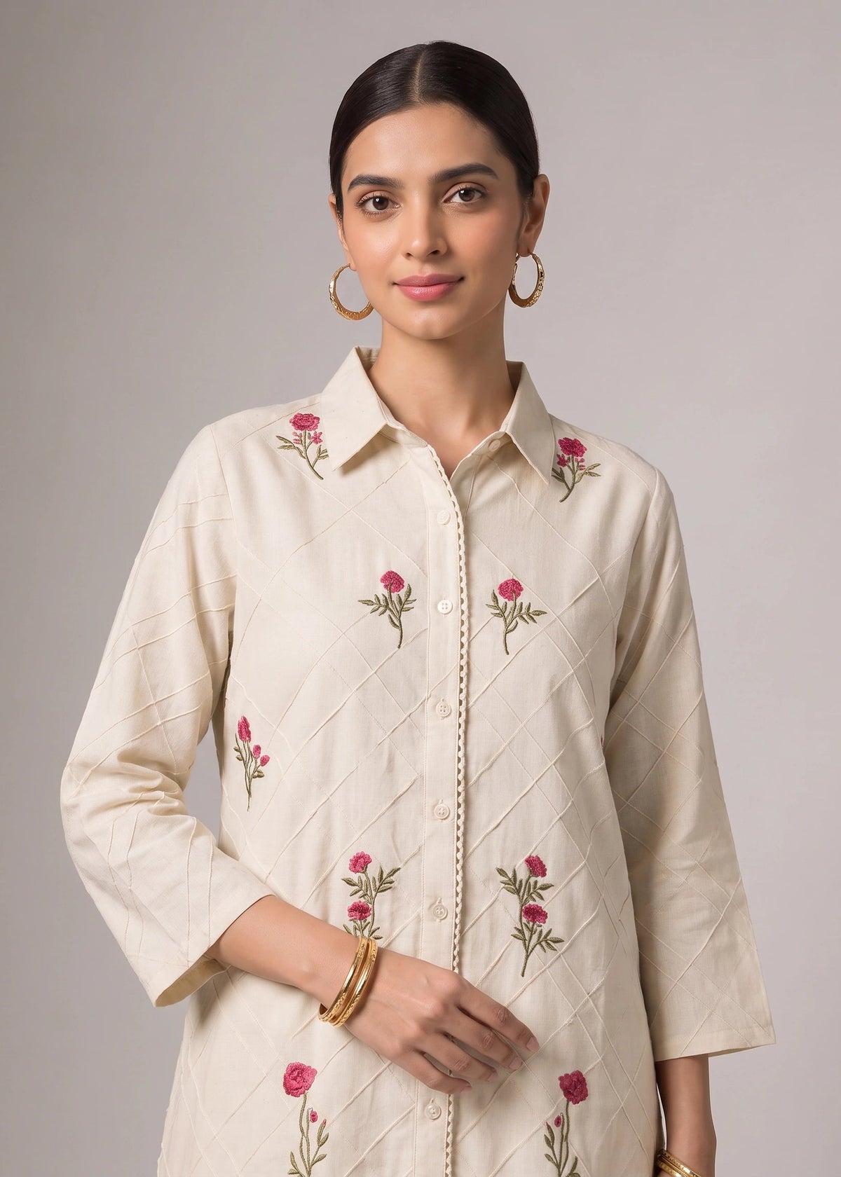 A woman modeling a cream-colored long-sleeve button-up shirt with pink floral embroidery and subtle diagonal quilted stitching, set against a solid gray studio background.