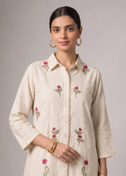 A woman modeling a cream-colored long-sleeve button-up shirt with pink floral embroidery and subtle diagonal quilted stitching, set against a solid gray studio background.