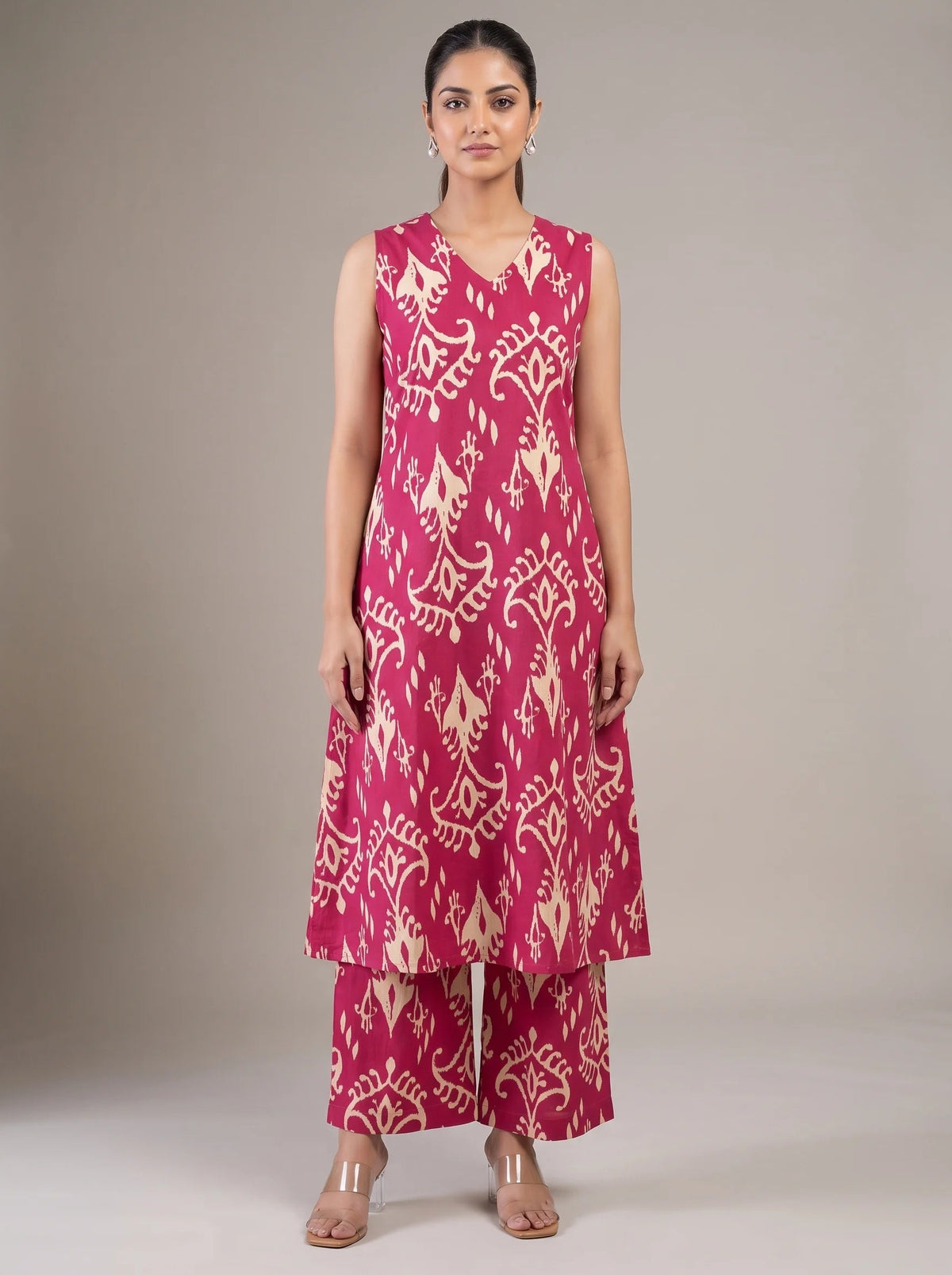 A woman modeling a sleeveless magenta pink and beige printed kurta set with matching wide-leg pants, standing against a neutral gray studio background.