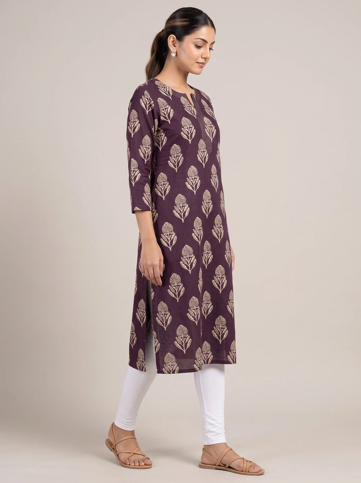 A woman modeling a three-quarter sleeve kurta in deep purple with a beige leaf block print, paired with white leggings and tan flat sandals, set against a plain beige studio background.