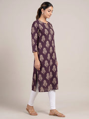 A woman modeling a three-quarter sleeve kurta in deep purple with a beige leaf block print, paired with white leggings and tan flat sandals, set against a plain beige studio background.