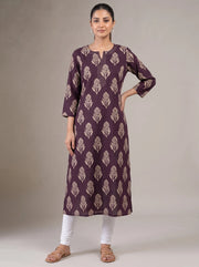 A woman modeling a three-quarter sleeve kurta in deep purple with a beige leaf block print, paired with white pants and brown sandals, against a neutral studio backdrop.