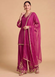 A woman modeling a magenta pink traditional Indian outfit with matching sheer dupatta, featuring gold embroidery and border details, set against a solid beige studio background.