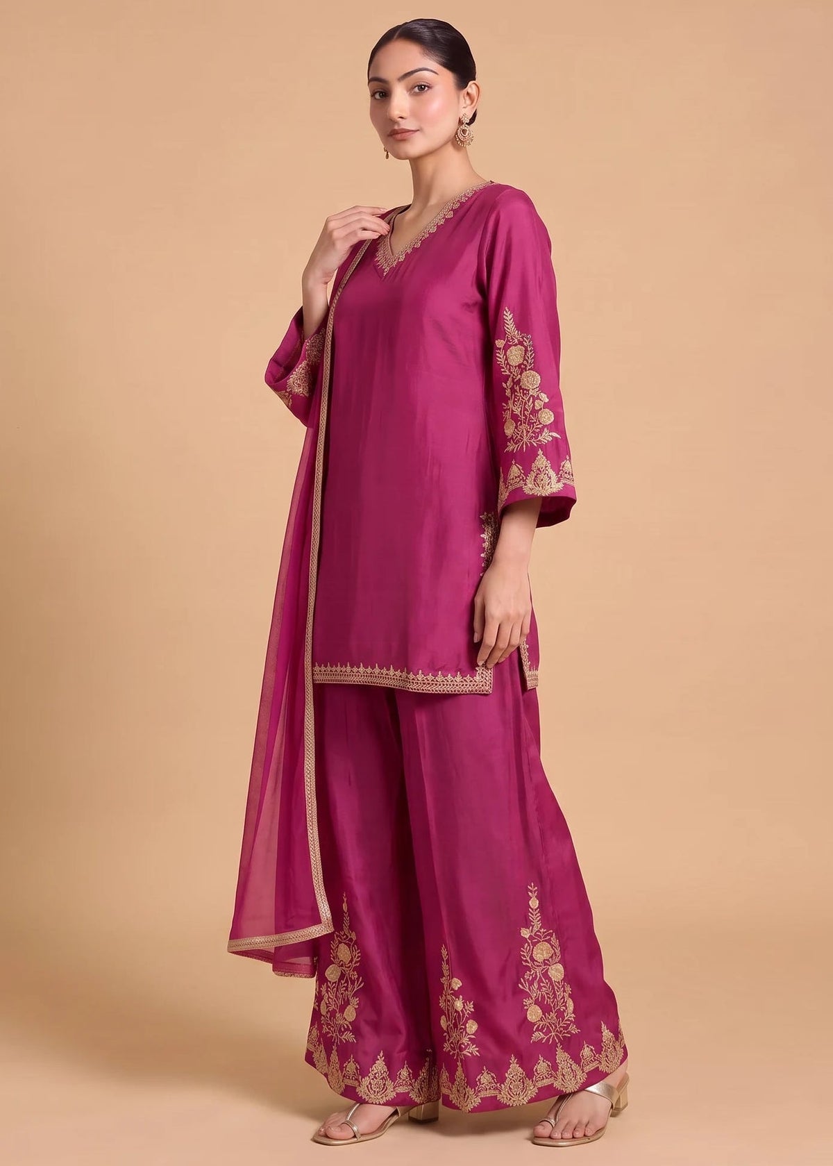 A woman modeling a magenta pink traditional Indian salwar kameez suit with gold embroidery, featuring a kurta, wide-leg pants, and a matching dupatta, set against a solid beige background.