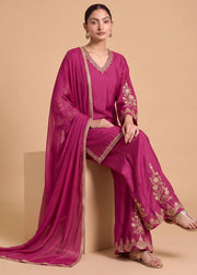 A woman models a magenta pink traditional three-piece outfit with gold embroidery, consisting of a V-neck kurta, wide-leg pants, and a matching dupatta, posed against a solid beige background.