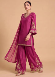 A woman models a magenta pink traditional Indian salwar kameez suit with gold embroidery, featuring a kurta, wide-leg trousers, and a matching dupatta, set against a solid beige background.