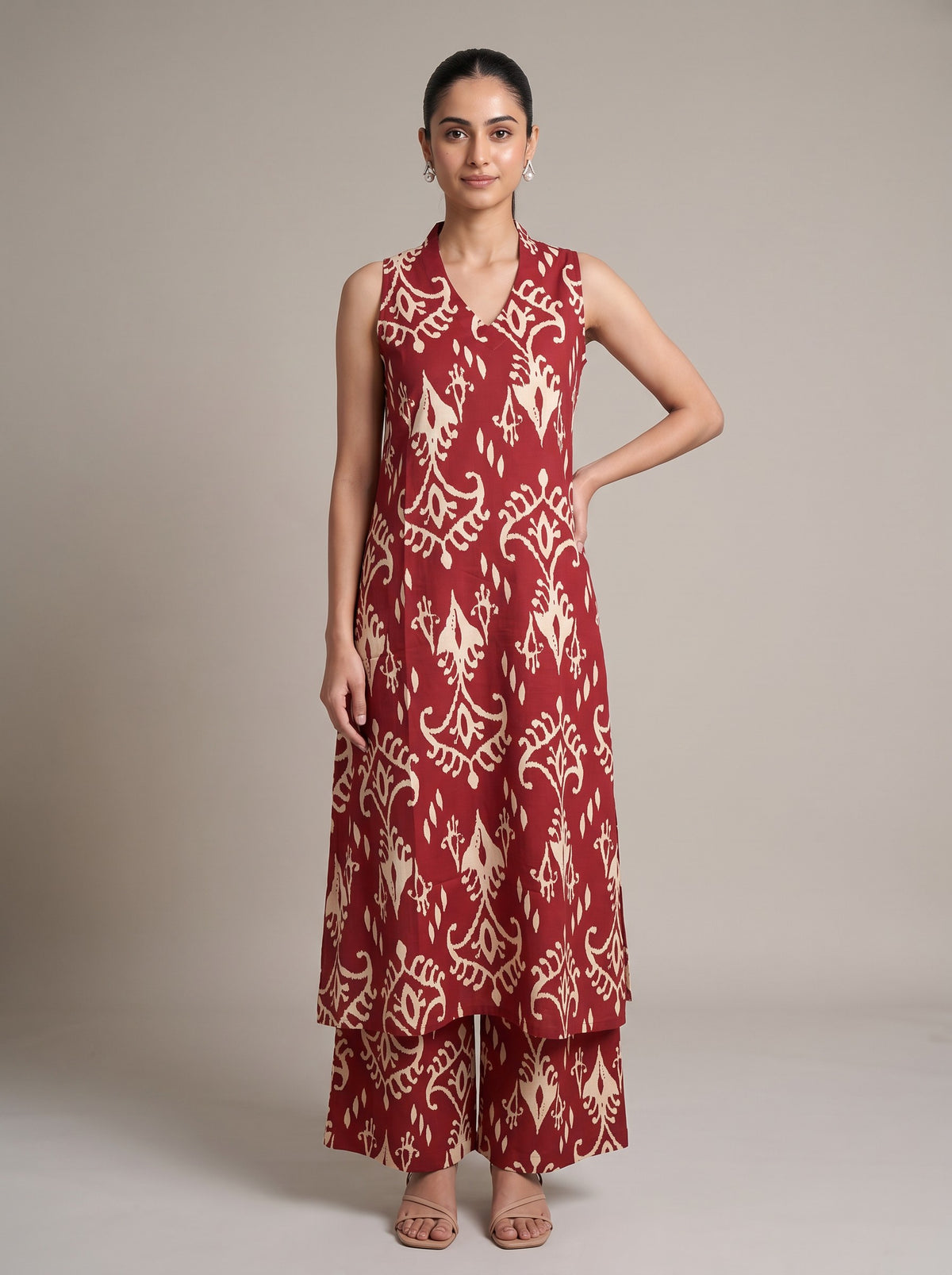 A woman modeling a sleeveless red and beige printed kurta with matching wide-leg pants against a plain beige studio background.