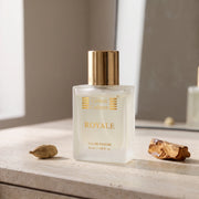A bottle of Cotton Culture ROYALE Eau de Parfum with a gold cap and gold lettering on a frosted glass bottle, styled on a stone surface with a cardamom and a piece of wood.