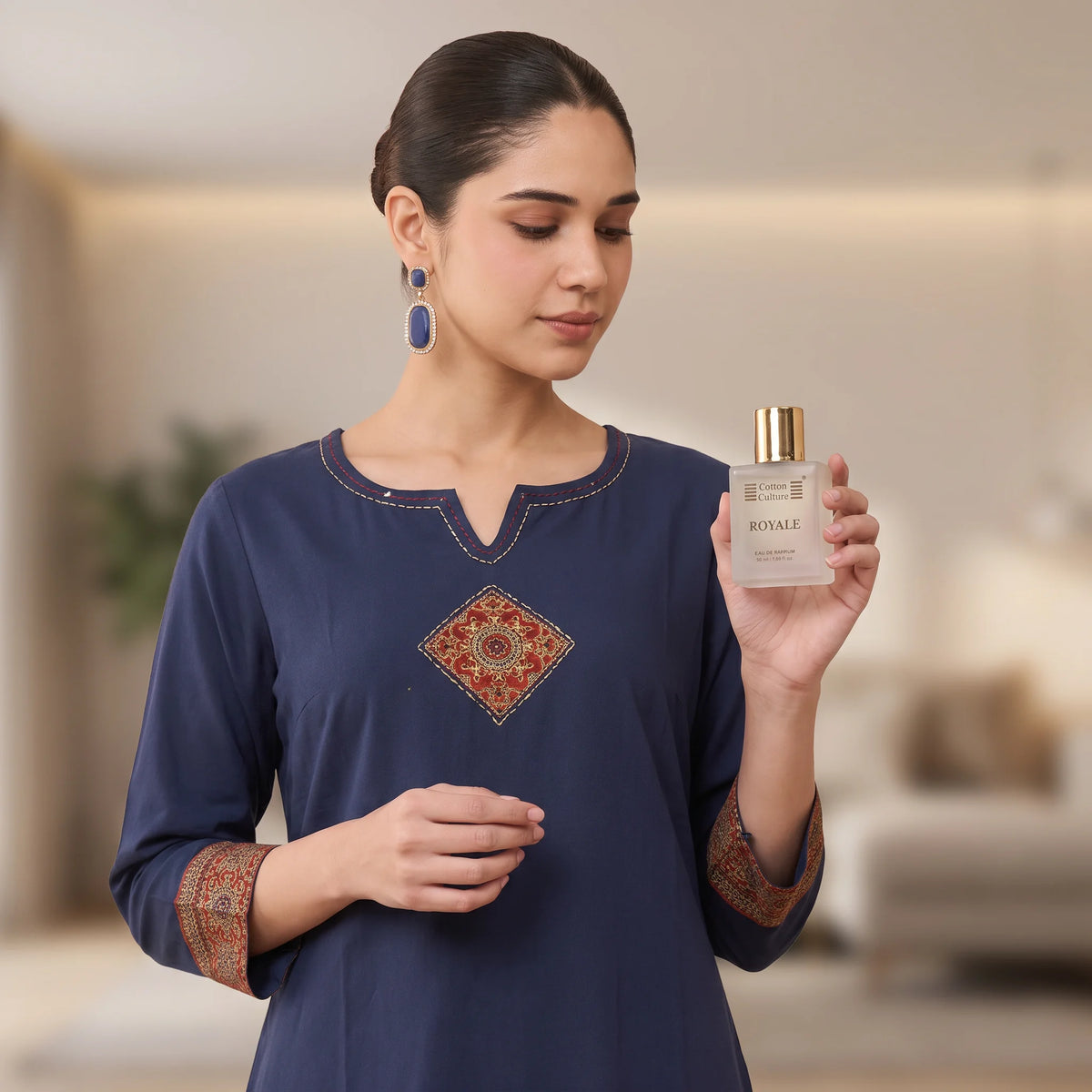 A woman wearing a navy blue embroidered kurta holds up a clear glass bottle of ROYALZ perfume. The kurta features a V-neckline with gold embroidery, a central diamond-shaped mandala embroidery, and patterned gold and red cuffs.