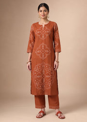 A woman modeling a rust-orange kurta set with white printed mandala patterns, paired with matching pants, against a solid beige studio background.