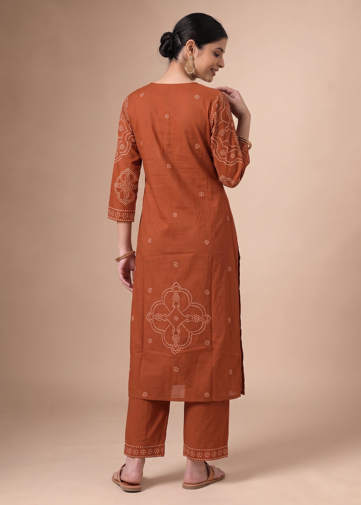 A woman modeling a rust-orange kurta and trouser set with beige embroidery, shown from the back against a solid beige studio backdrop.