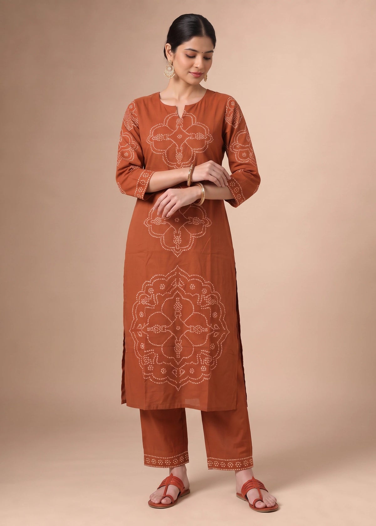 A woman modeling a rust-orange kurta set with white printed mandala patterns, paired with matching pants and brown sandals, against a solid beige studio background.