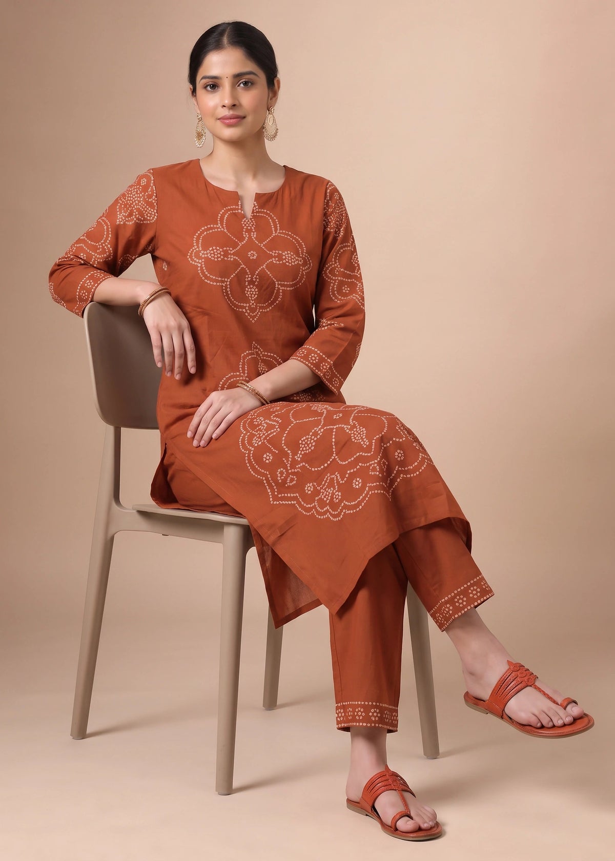 A woman seated on a light beige chair wearing a rust-orange kurta set with white embroidered motifs and matching pants, paired with matching flat sandals, against a solid beige studio background.