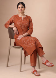 A woman seated on a light beige chair wearing a rust-orange kurta set with white embroidered motifs and matching pants, paired with matching flat sandals, against a solid beige studio background.