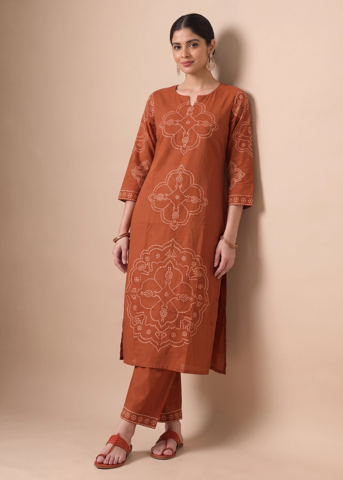 A woman modeling a rust-orange kurta set with beige floral embroidery, paired with matching pants and brown sandals, standing against a solid beige studio backdrop.