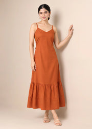 A woman modeling a rust-orange sleeveless maxi dress with thin straps, a V-neckline, and a tiered ruffle hem, standing against a solid beige studio background.