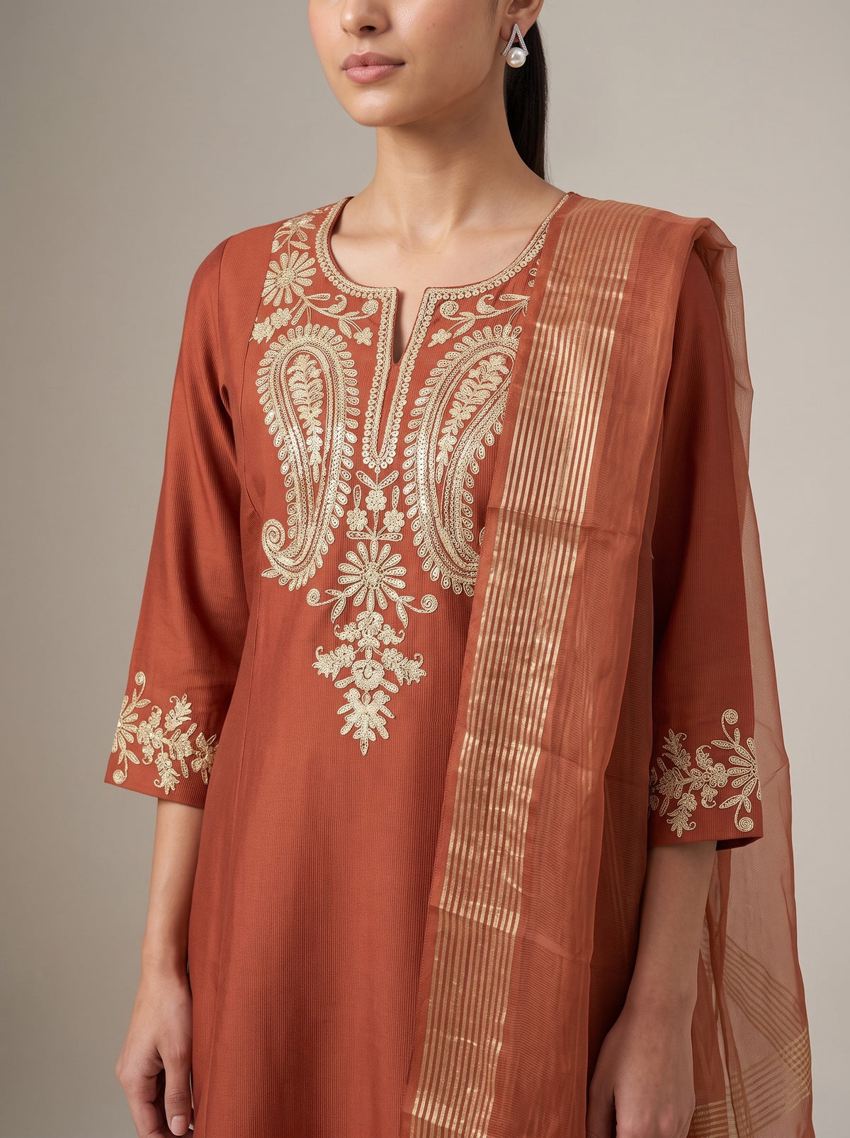 A woman wearing a rust-orange kurta with intricate gold paisley embroidery on the chest and sleeve cuffs, paired with a matching sheer dupatta featuring a gold striped border.