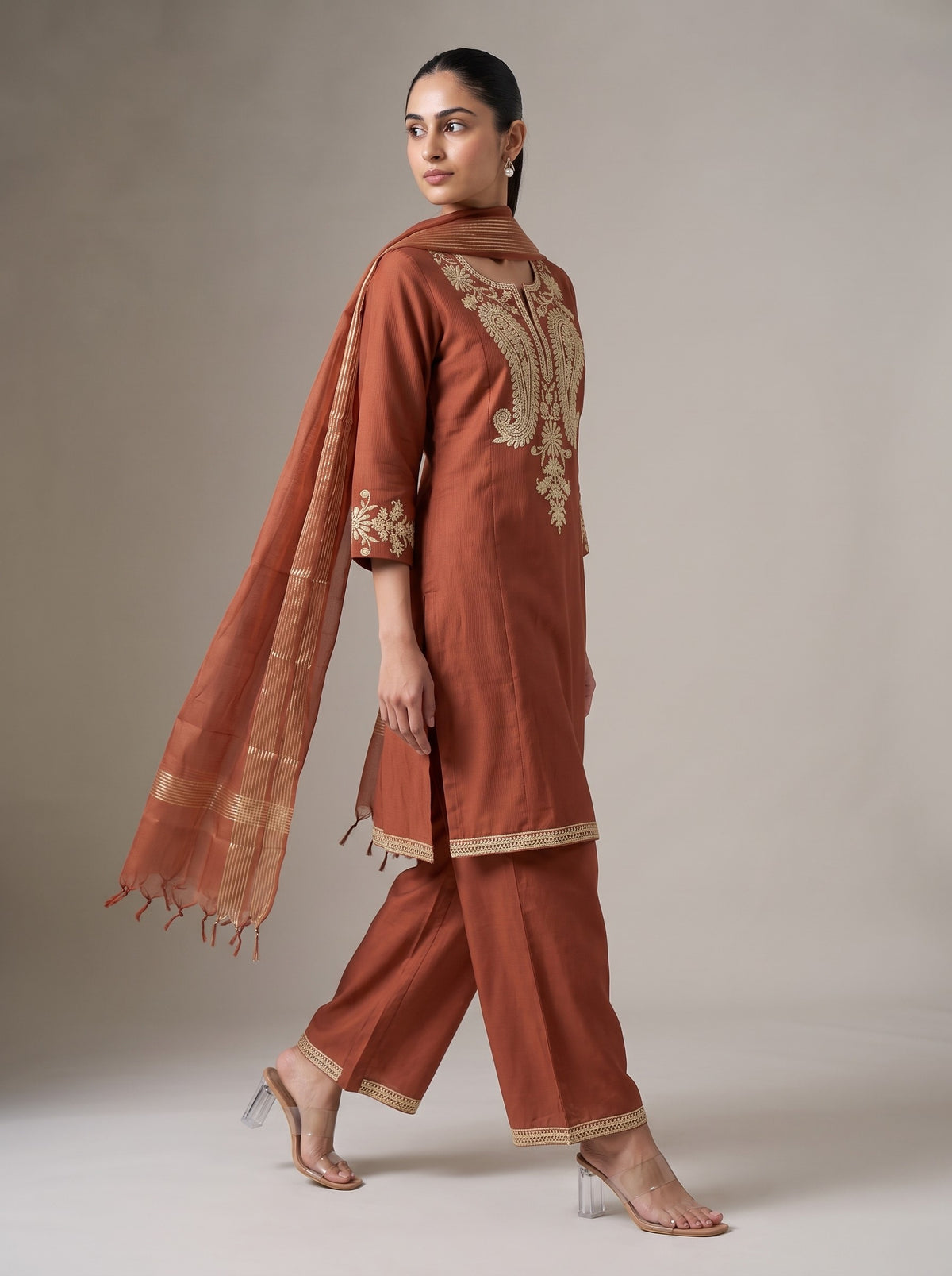 A woman modeling a rust-orange traditional Indian salwar kameez suit with gold embroidery on the back and cuffs, paired with matching wide-leg trousers and a sheer dupatta, set against a neutral studio background.