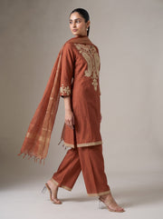 A woman modeling a rust-orange traditional Indian salwar kameez suit with gold embroidery on the back and cuffs, paired with matching wide-leg trousers and a sheer dupatta, set against a neutral studio background.