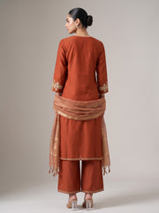 A woman shown from the back wearing a rust-orange traditional outfit with matching pants and a sheer dupatta, photographed against a plain light gray studio background.