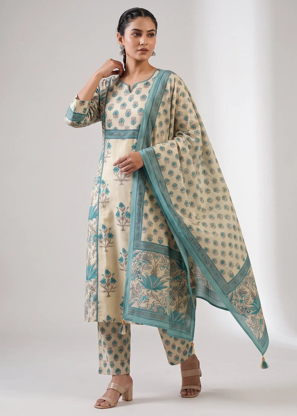 A woman modeling a traditional Indian salwar kameez suit set in cream with teal blue floral prints and borders, including a matching dupatta, against a neutral studio background.