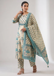 A woman modeling a traditional Indian salwar kameez suit set in cream with teal blue floral prints and borders, including a matching dupatta, against a neutral studio background.