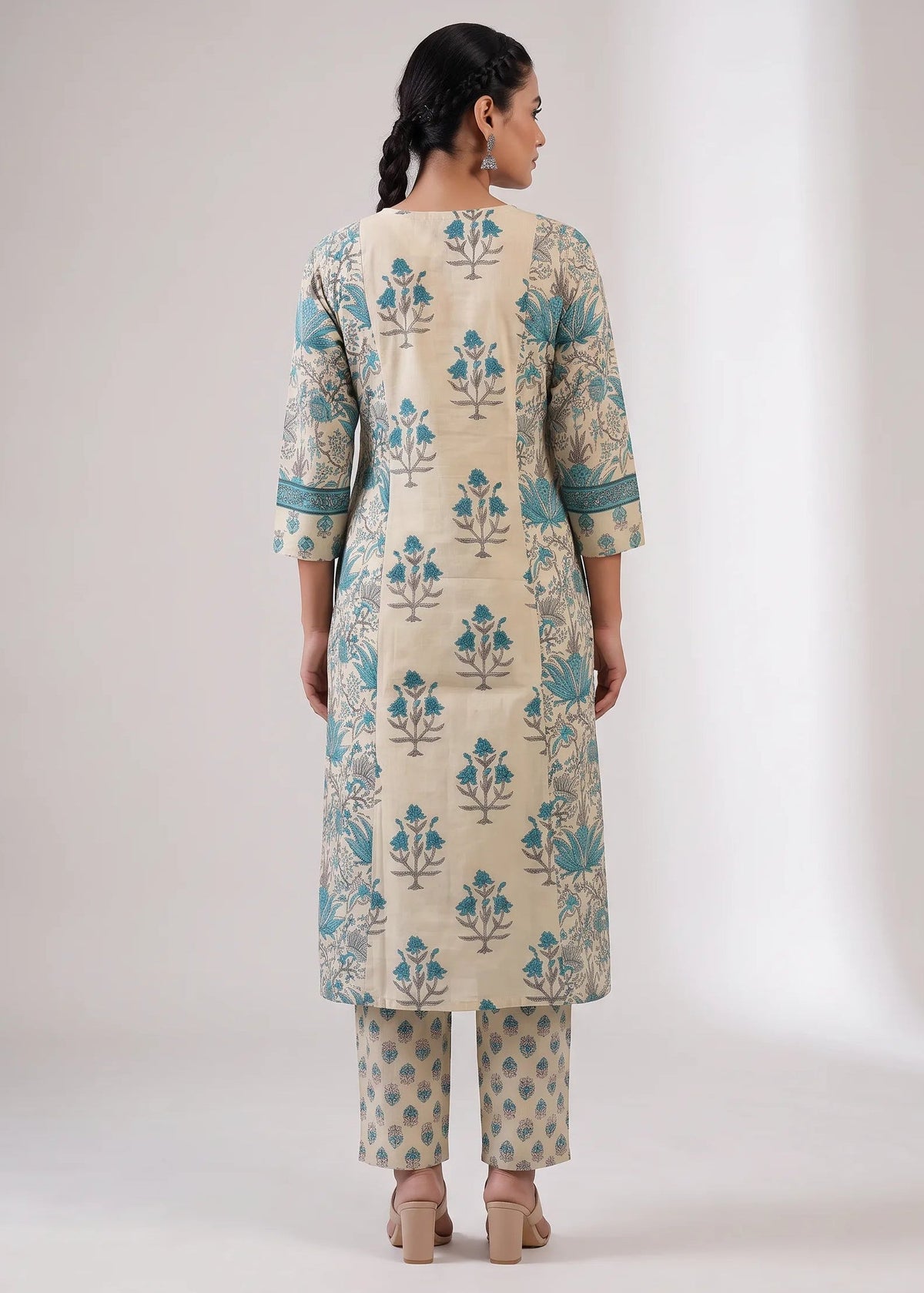 A woman viewed from the back wearing a cream-colored kurta with teal blue floral block prints and matching printed pants, set against a plain light gray studio background.