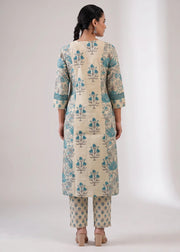 A woman viewed from the back wearing a cream-colored kurta with teal blue floral block prints and matching printed pants, set against a plain light gray studio background.