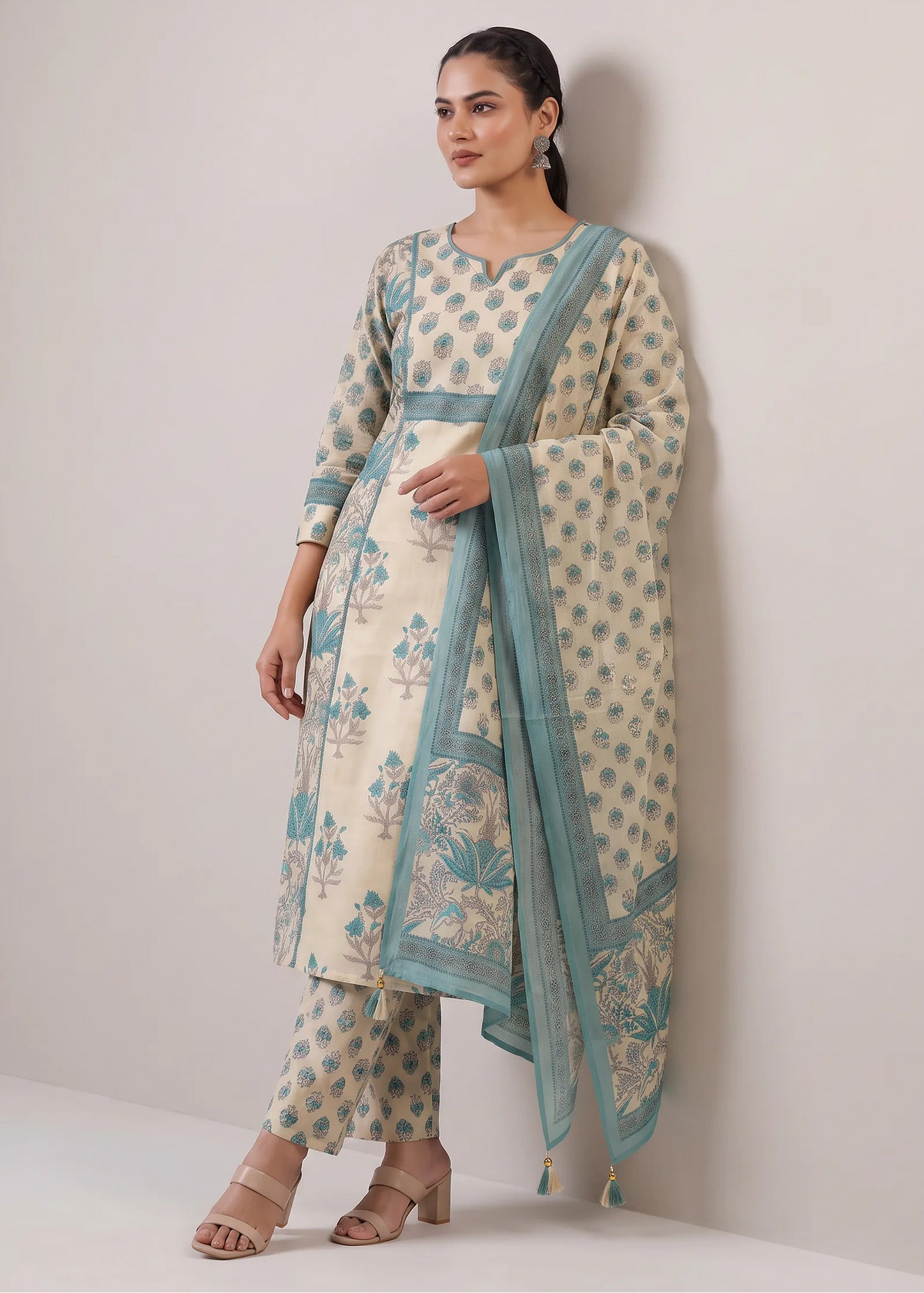 A woman models a traditional Indian salwar kameez set consisting of a cream-colored kurta and pants with teal blue floral prints and borders, paired with a matching dupatta, against a plain light gray wall.