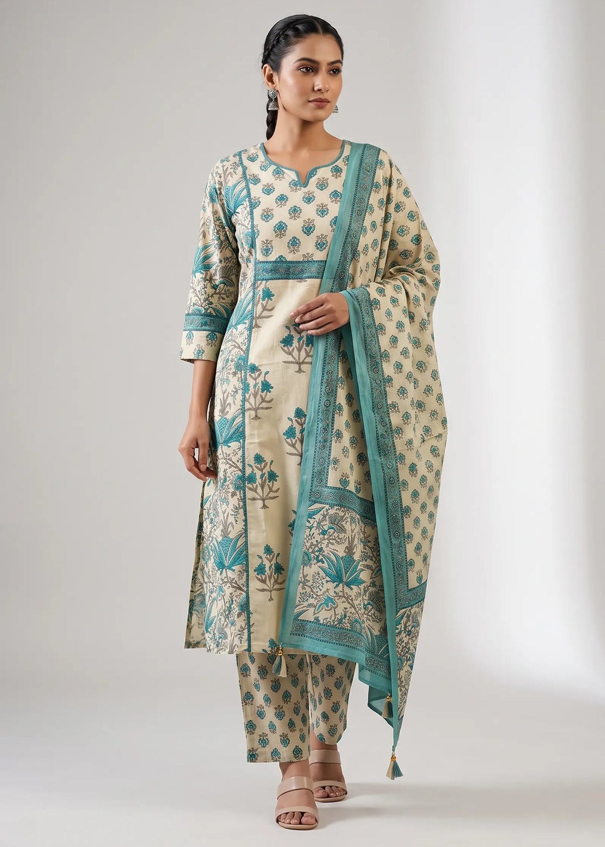 A woman modeling a traditional Indian salwar kameez suit set in beige with teal blue floral block prints and borders, paired with matching trousers and a dupatta, against a neutral studio background.
