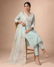A woman models a light blue traditional Indian salwar kameez suit with a white embroidered yoke, matching trousers, and a sheer white dupatta with a gold border, seated on a wooden chair against a solid beige background.