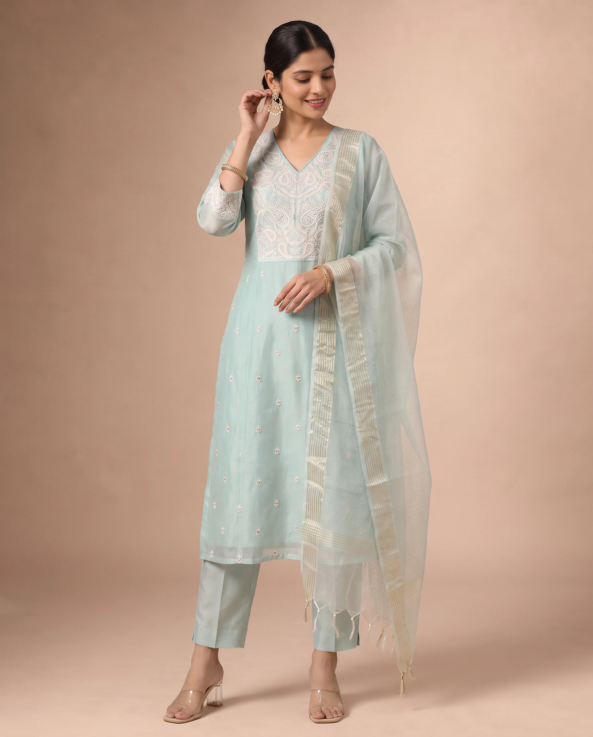 A woman modeling a light blue-green traditional Indian salwar kameez suit with white embroidery, matching trousers, and a sheer white dupatta, set against a solid beige studio background.